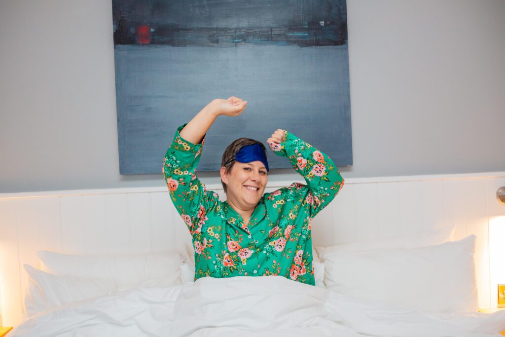 A woman in her forties in green pyjamas sits up and stretches in bed in a hotel room. She has a sleep mask on her head.