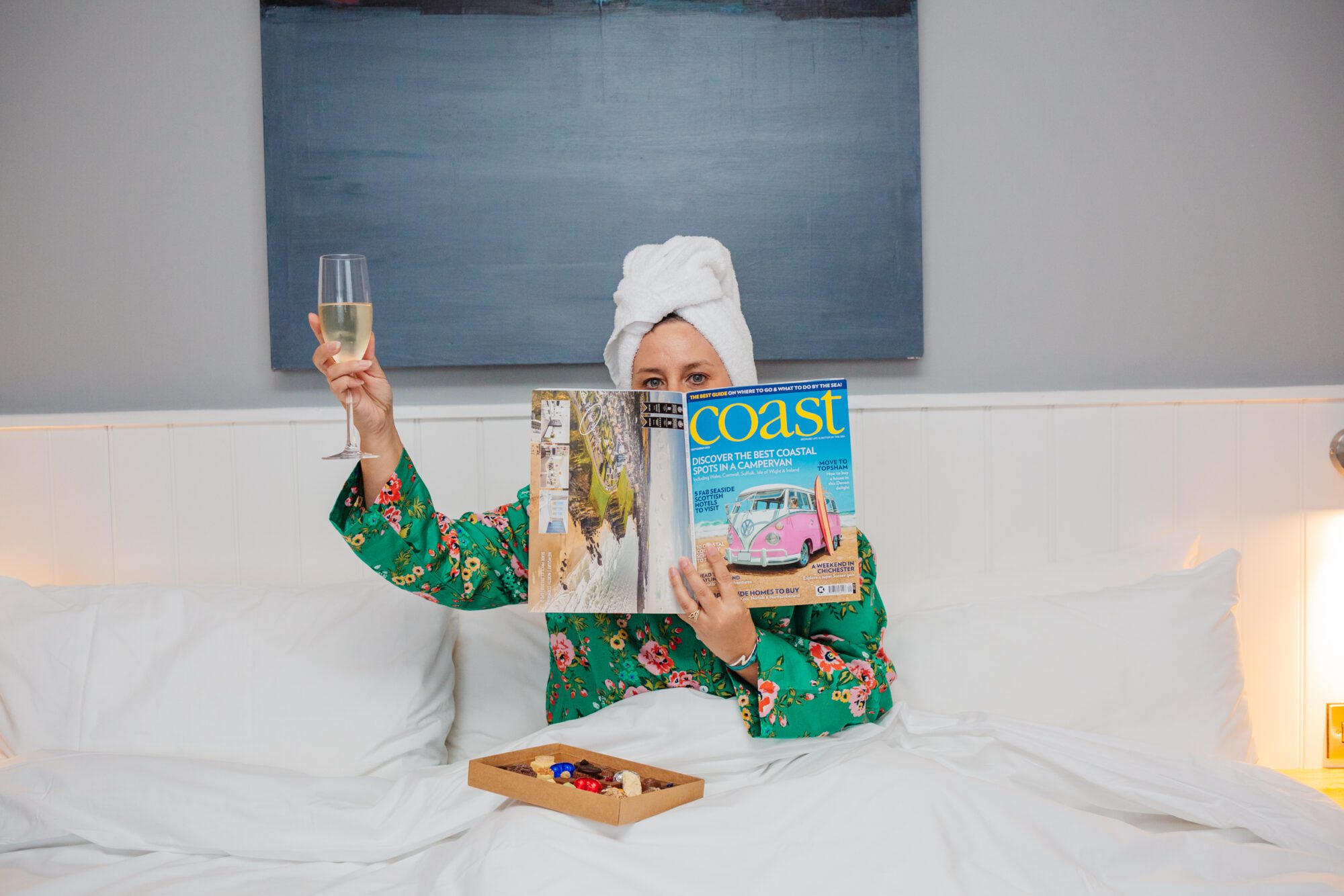A woman in bed in silky pyjamas holding a glass of fizz in one hand and a magazine in the other covering her face