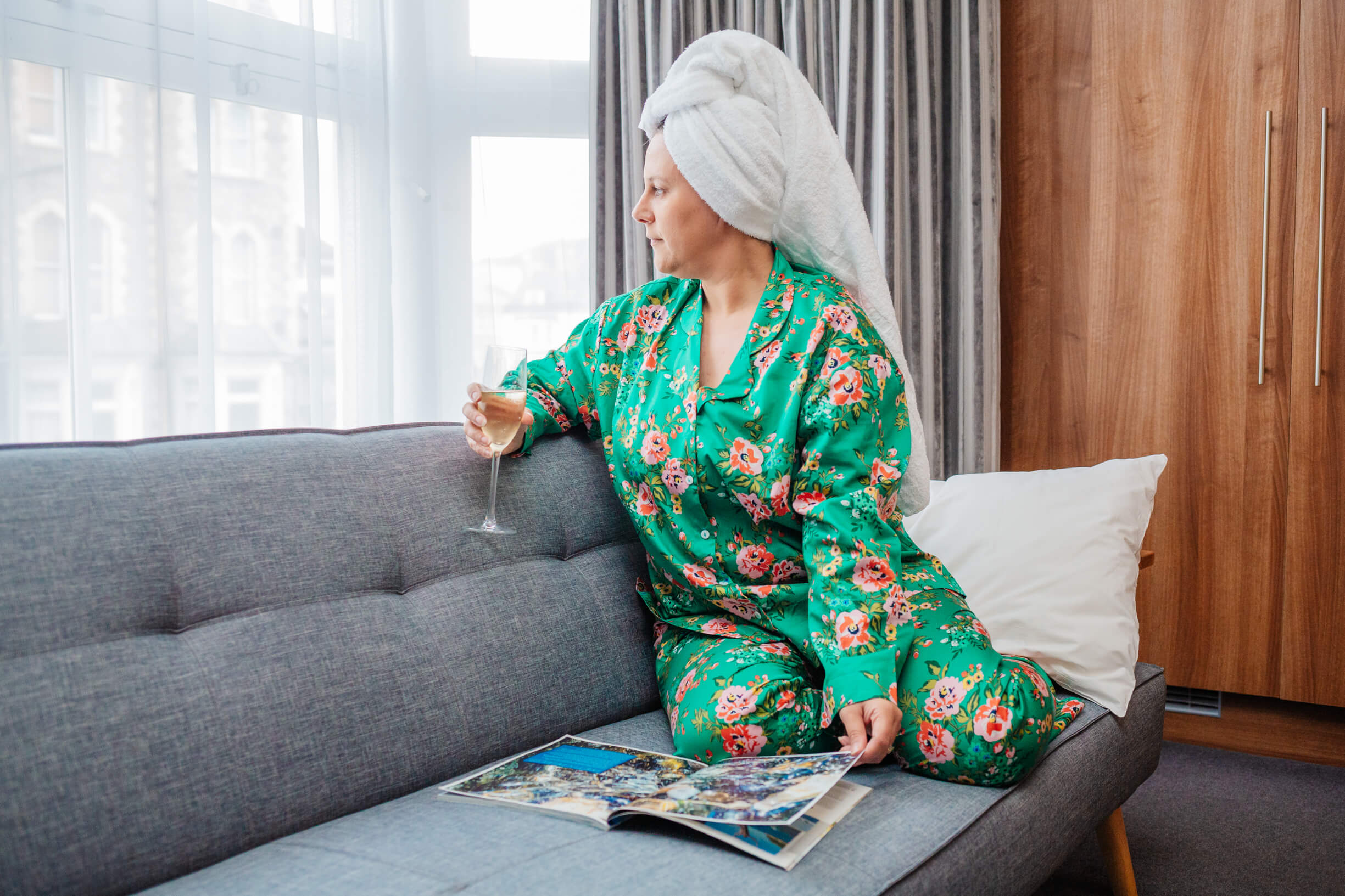 A woman in her forties sits on a sofa in a hotel room reading a magazine and wearing silky pyjamas