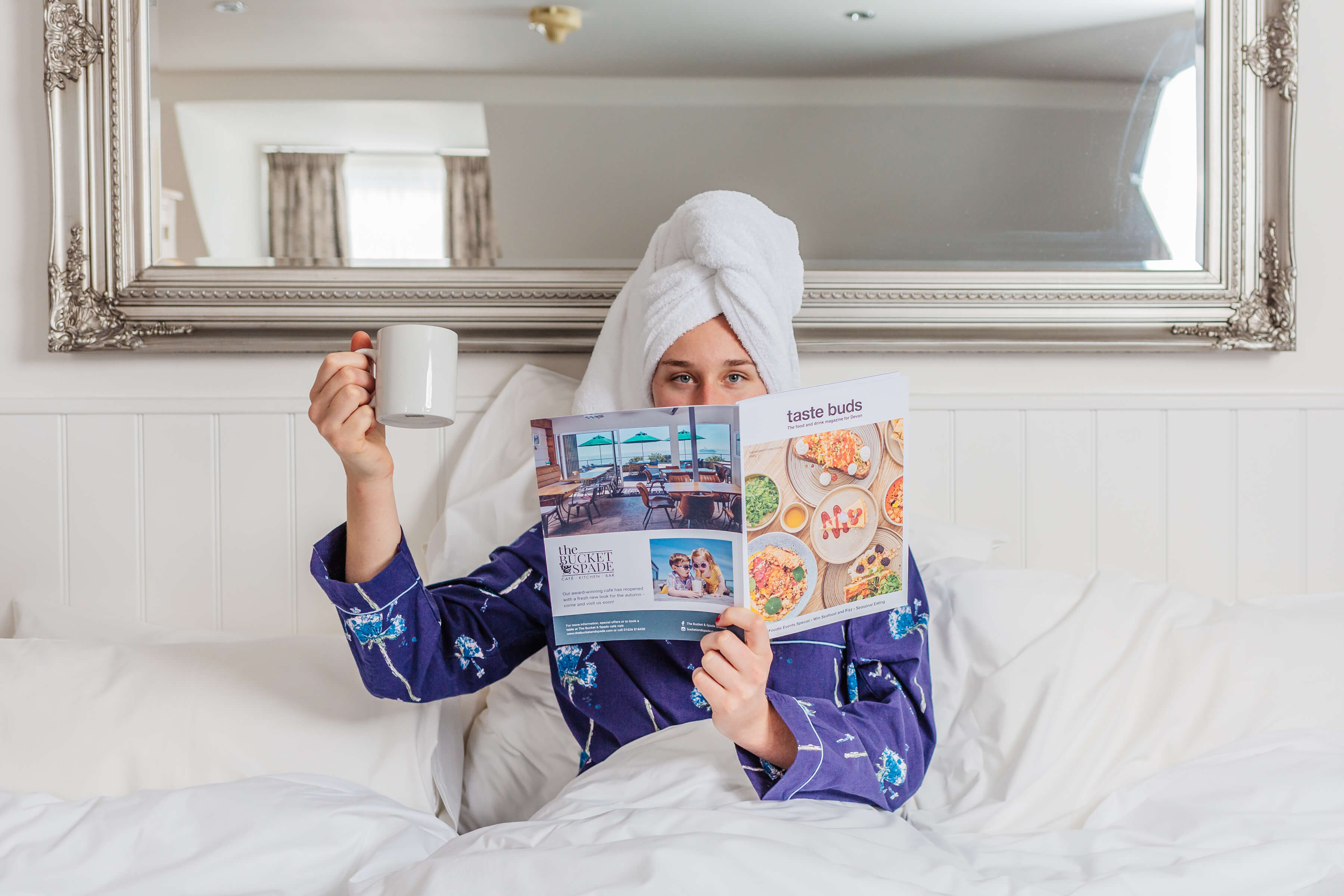 A woman in a hotel bed holding a magazine in front of her face and a mug of tea in her hand